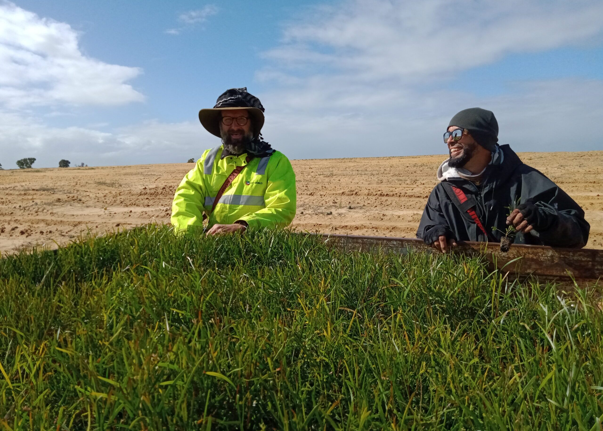 Two men in hats and glasses standing behind a trailer full of seedlings